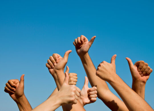 Group of people giving thumbs up over blue sky